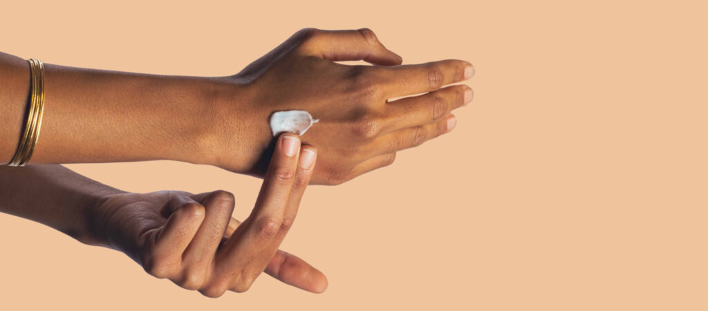 A person applies lotion to the back of their hand with a finger against a beige background.