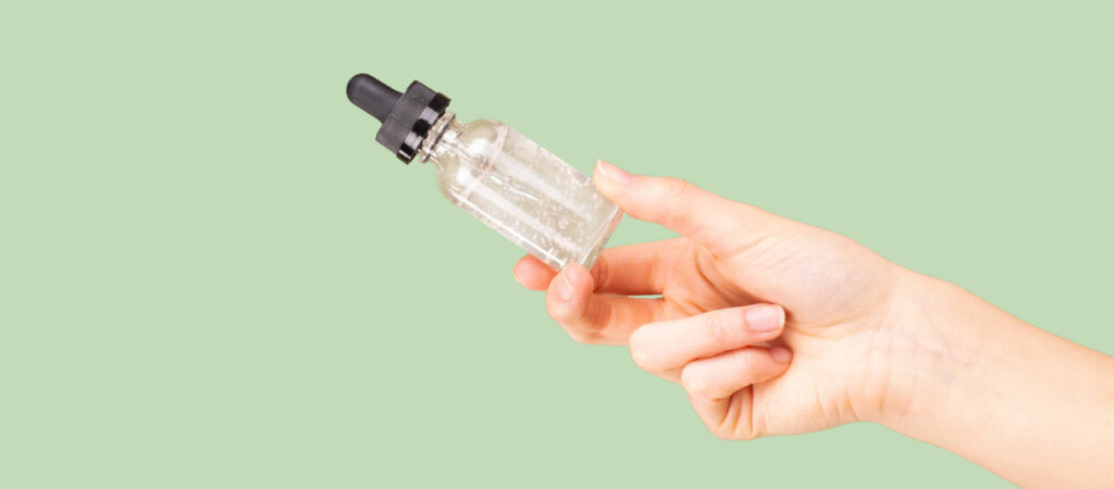 A hand holding a clear glass dropper bottle with a black cap, set against a light green background.