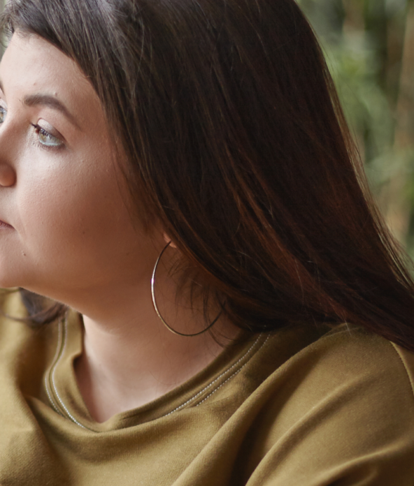 A woman with long brown hair and hoop earrings gazes out a window. She is wearing a mustard-colored sweatshirt, and greenery is visible in the blurred background.