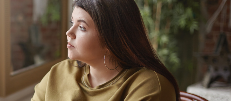 A woman with long brown hair and hoop earrings gazes out a window. She is wearing a mustard-colored sweatshirt, and greenery is visible in the blurred background.