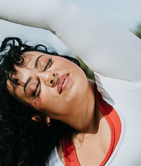 Person stretching outdoors with eyes closed, wearing a white long-sleeved top. Trees and sky in the background.