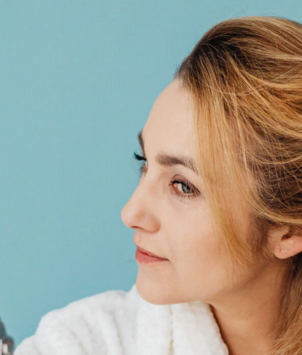 A woman brushes her hair while looking into a mirror against a blue background.