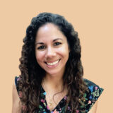 A person with long curly hair smiles, wearing a floral-patterned top and gold necklace, against a beige background.