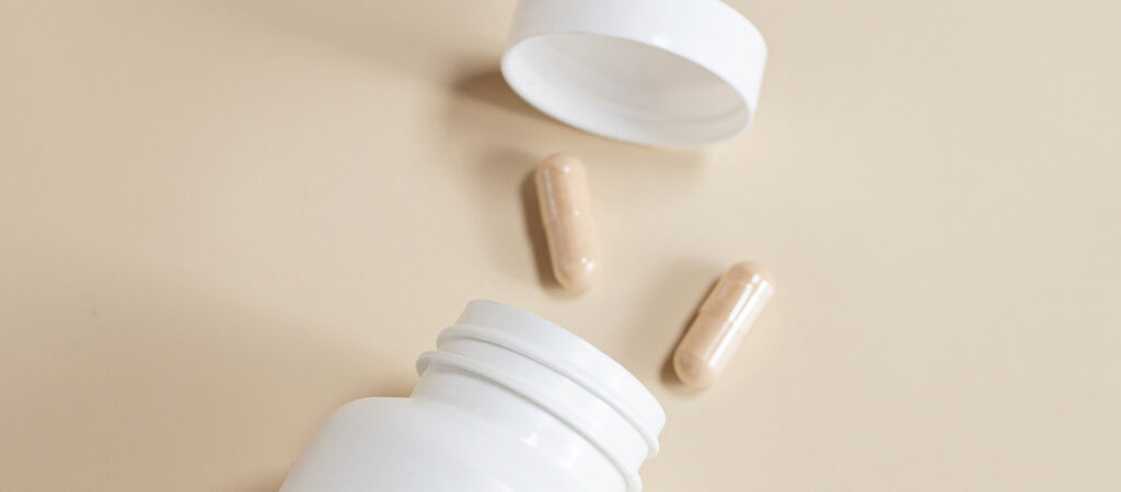 Open white pill bottle with three beige capsules and a lid on a light surface.