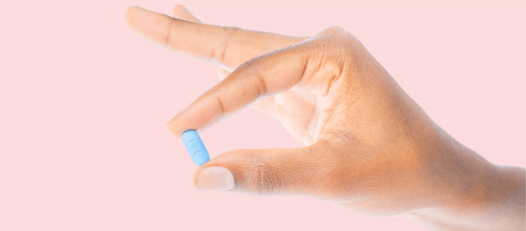 A hand holds a small blue pill between the thumb and index finger against a pink background.