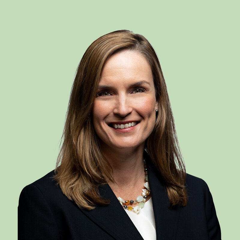 A woman with long brown hair wearing a dark blazer and beaded necklace smiles against a light green background.