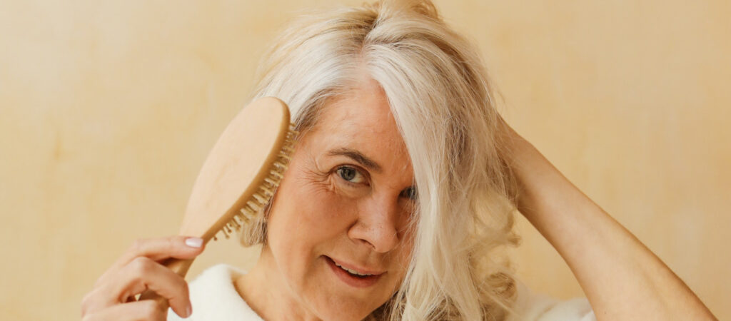 Person with long gray hair brushes their hair, smiling, against a beige background.