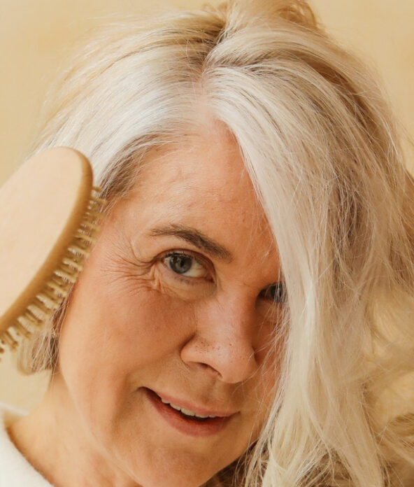 Person with long gray hair brushes their hair, smiling, against a beige background.