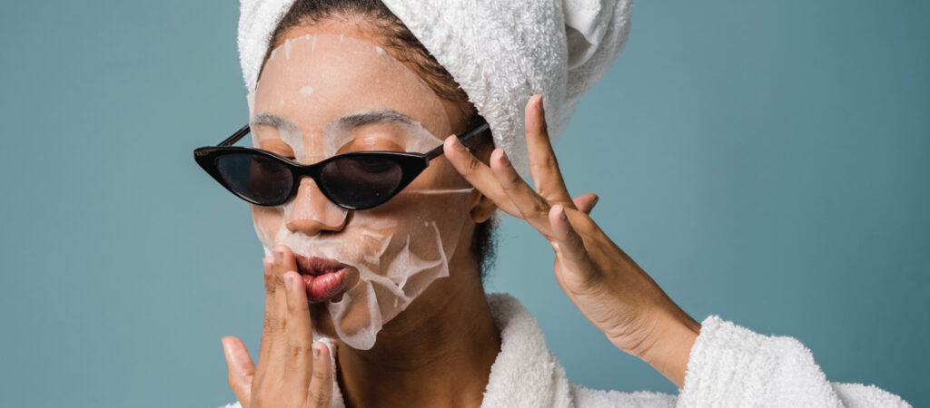 Person wearing a face mask, cat-eye sunglasses, and a towel on their head, with one hand touching their lips against a blue background.