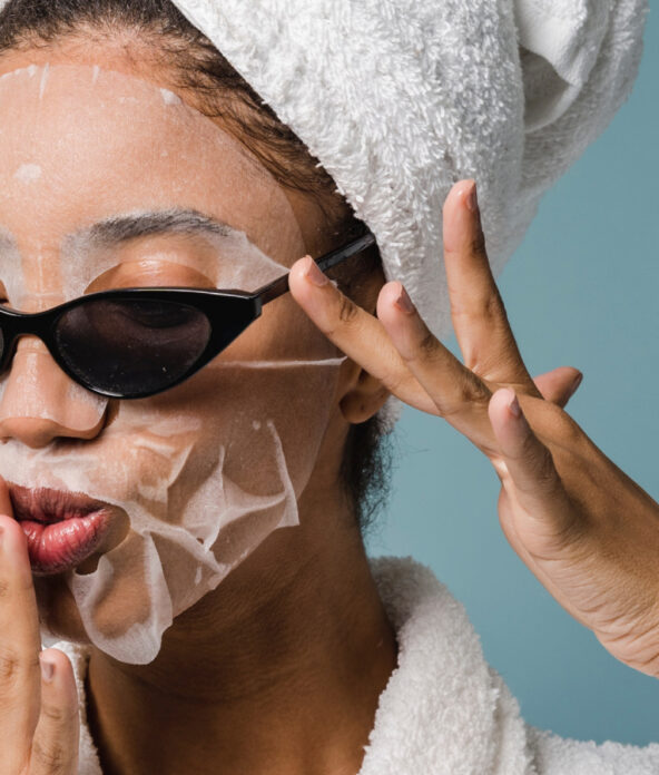 Person wearing a face mask, cat-eye sunglasses, and a towel on their head, with one hand touching their lips against a blue background.