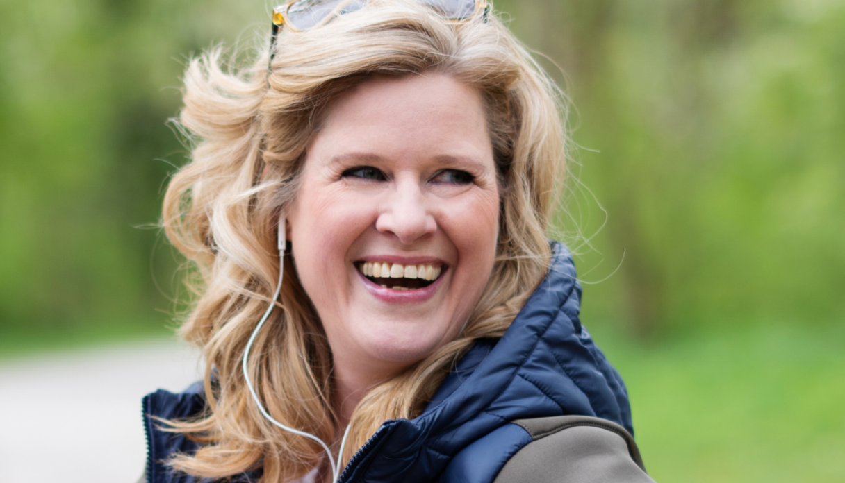 Person with wavy blonde hair, wearing a blue vest, smiling outdoors with greenery in the background.