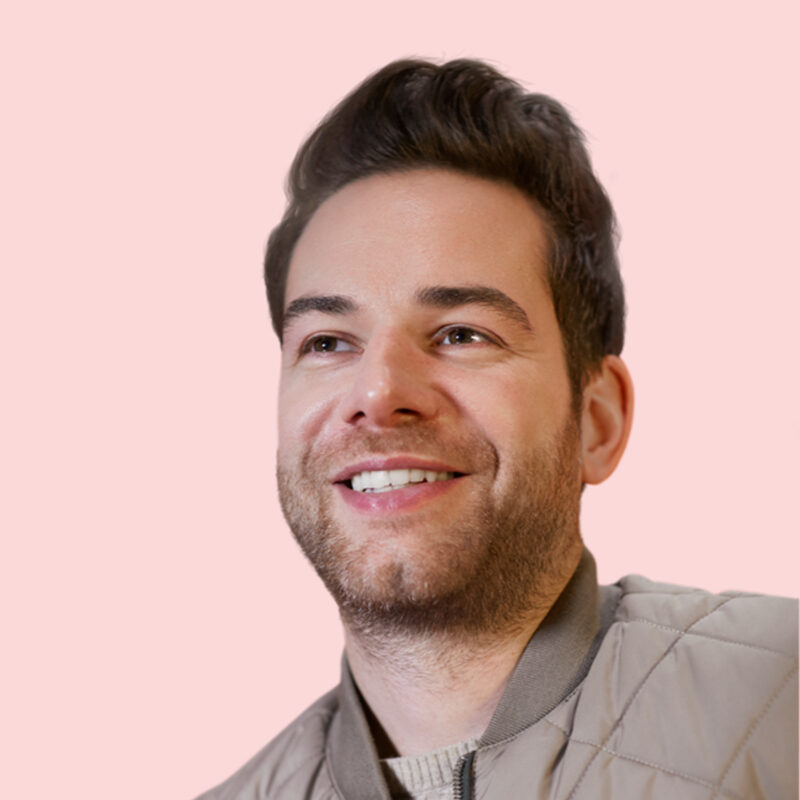 Smiling man with short dark hair and a beard, wearing a light brown jacket, against a pale pink background.