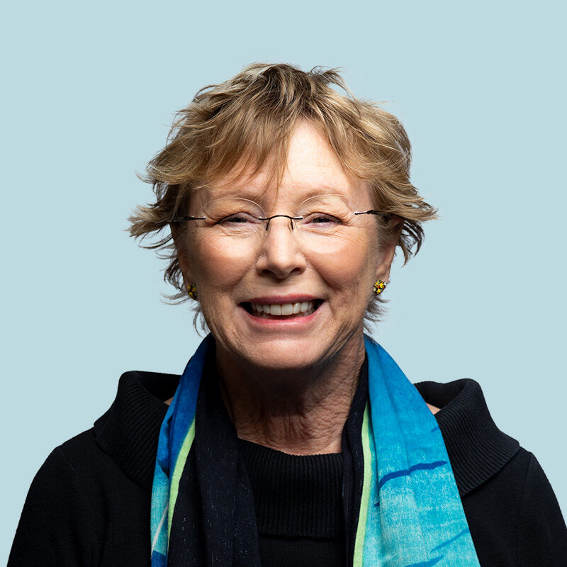 An older woman with glasses and short hair smiles, wearing a black top and blue scarf, against a light blue background.