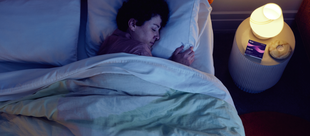 A person sleeps in bed beside a nightstand with a glowing lamp and a packet of sleep aid tablets.