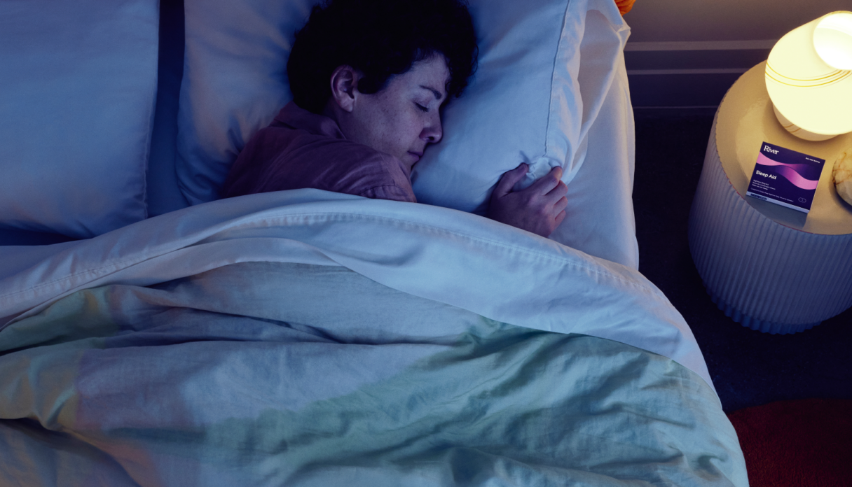 A person sleeps in bed beside a nightstand with a glowing lamp and a packet of sleep aid tablets.