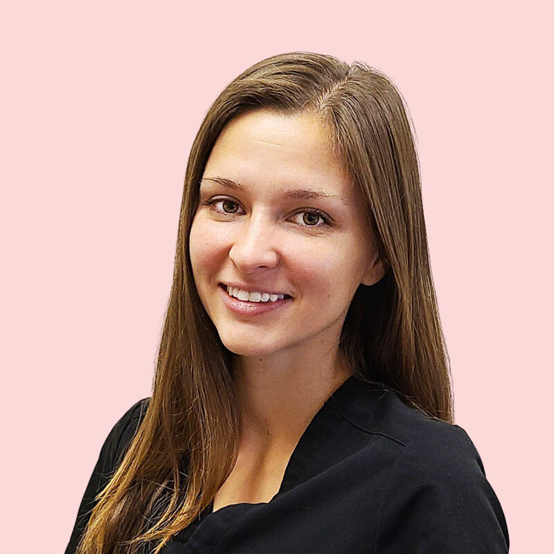 A person with long brown hair wearing a black top smiles against a pink background.