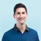 Man in a blue collared shirt smiling against a light blue background.